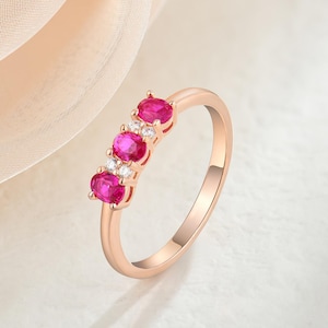 Può includere: Anello in oro rosa con tre pietre rosse ovali e piccoli accenti chiari. L'anello ha una fascia sottile e lucida. Le pietre preziose sono disposte in fila, offrendo un look classico ed elegante. Un gioiello.
