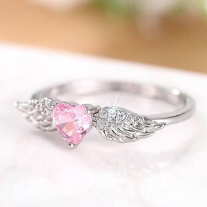May include: A silver ring featuring a pink heart-shaped gemstone, flanked by delicate wing designs. The ring has a polished finish and is set against a soft, blurred background, highlighting the ring's intricate details and sparkle.
