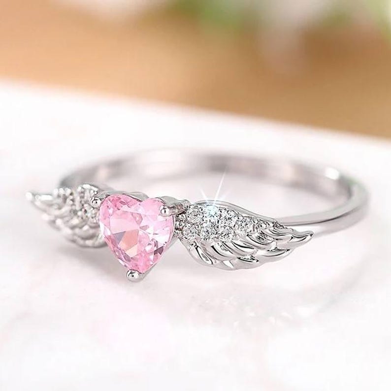 May include: A silver ring featuring a pink heart-shaped gemstone, flanked by delicate wing designs. The ring has a polished finish and is set against a soft, blurred background, highlighting the ring's intricate details and sparkle.