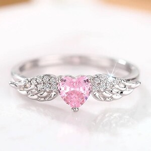 May include: A silver ring featuring a pink heart-shaped gemstone. The ring has small, sparkling accents and wing-shaped details on either side of the heart. The ring is set against a soft, blurred background.