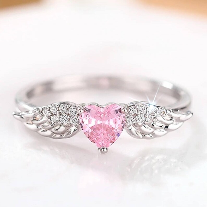 May include: A silver ring featuring a pink heart-shaped gemstone. The ring has small, sparkling accents and wing-shaped details on either side of the heart. The ring is set against a soft, blurred background.
