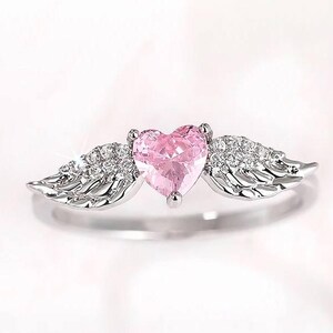 May include: A silver ring featuring a pink heart-shaped gemstone, flanked by detailed wing designs encrusted with small, sparkling stones. The ring has a polished finish and a delicate, romantic aesthetic.