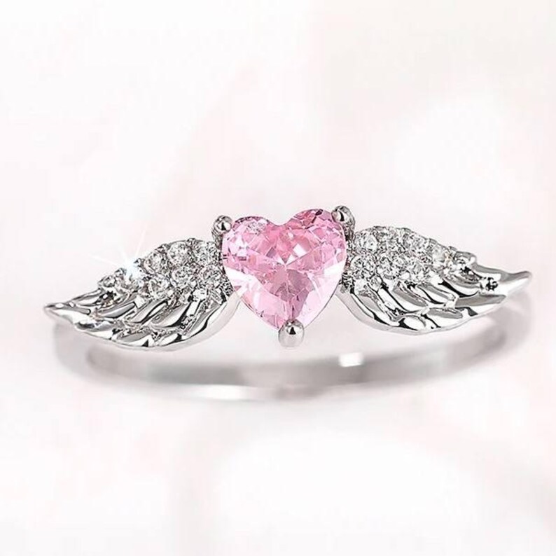 May include: A silver ring featuring a pink heart-shaped gemstone, flanked by detailed wing designs encrusted with small, sparkling stones. The ring has a polished finish and a delicate, romantic aesthetic.