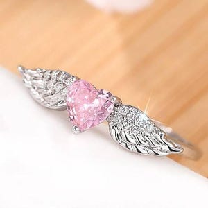 May include: A silver ring featuring a pink heart-shaped gemstone, flanked by detailed wing designs. The ring's band is thin and polished, with the wings and heart adding a decorative element. The ring is set against a soft, neutral background.