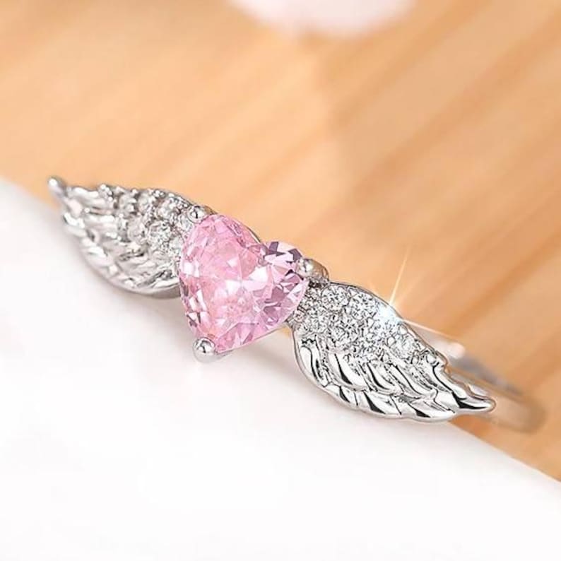 May include: A silver ring featuring a pink heart-shaped gemstone, flanked by detailed wing designs. The ring's band is thin and polished, with the wings and heart adding a decorative element. The ring is set against a soft, neutral background.