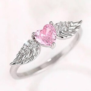 May include: A silver ring featuring a pink heart-shaped gemstone at the center, flanked by detailed wing designs. The wings are embellished with small, sparkling stones. The ring is set against a soft, white background.