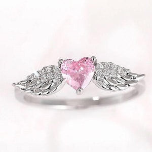 May include: A silver ring featuring a pink heart-shaped gemstone, flanked by two wings embellished with small, sparkling stones. The ring has a polished finish and is set against a soft, white background.