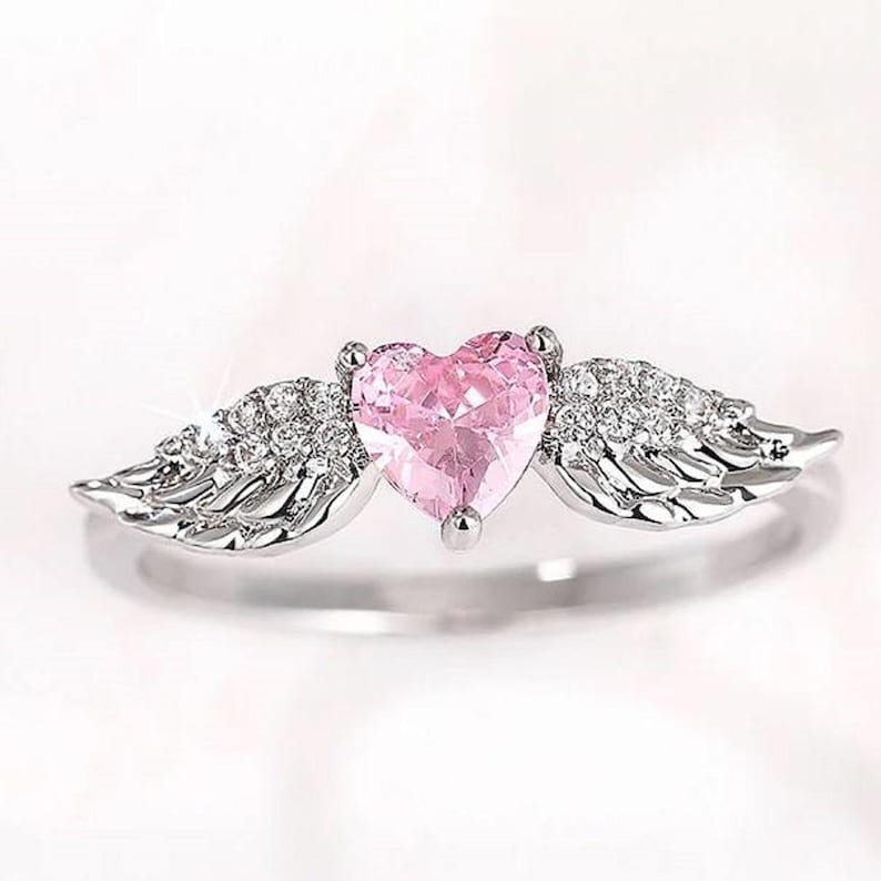 May include: A silver ring featuring a pink heart-shaped gemstone, flanked by two wings embellished with small, sparkling stones. The ring has a polished finish and is set against a soft, white background.