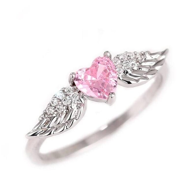 May include: A silver ring featuring a pink heart-shaped gemstone, flanked by detailed wing designs. The wings are embellished with small, clear gemstones. The ring is set against a white background, highlighting the jewelry's delicate features.
