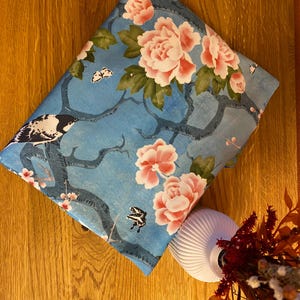 Könnte beinhalten: Ein gefalteter Stoff mit einem blauen Blumenmuster mit rosa Pfingstrosen, grünen Blättern und einem Vogeldesign. Der Stoff liegt auf einer Holzoberfläche. Das Design umfasst Schmetterlinge und Äste.