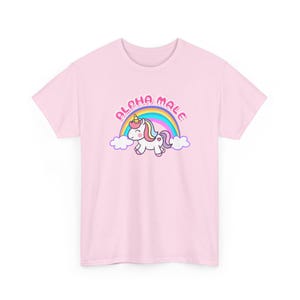 Op de afbeelding: Lichtroze T-shirt met een cartoon eenhoorn, een regenboog en de tekst "ALPHA MALE". De eenhoorn heeft een gele hoorn, roze manen en een paarse staart. De regenboog is veelkleurig.