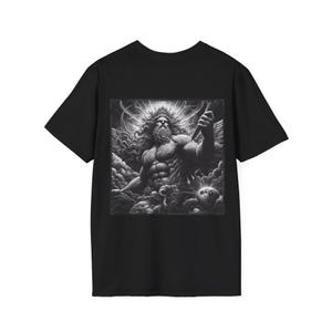 May include: Black t-shirt with a monochrome illustration of a muscular, bearded figure with a halo and wings, holding a weapon, set against a cloudy background. The graphic is centered on the shirt.