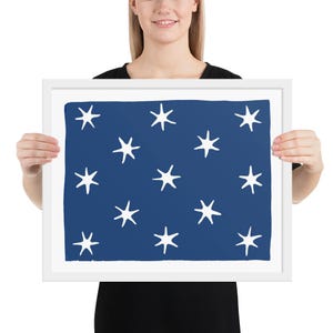 Peut inclure: Une impression d'art encadrée avec un fond bleu marine et des motifs d'éclats d'étoiles blancs. L'œuvre d'art est tenue par une personne, mettant en valeur le cadre blanc. Le design est simple et frappant.