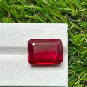 May include: A vibrant, deep red gemstone cut into a rectangular emerald shape with faceted edges. The stone displays a clear, transparent quality and is positioned on a white display stand, showcasing its polished surface and geometric symmetry.