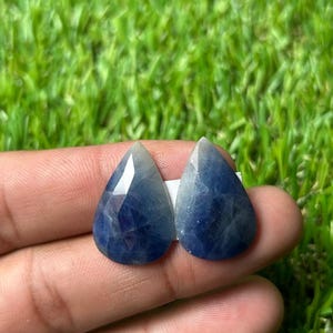 Puede incluir: Dos zafiros azules en forma de pera, facetados, con una apariencia translúcida e inclusiones internas visibles. Las piedras preciosas presentan una superficie pulida con cortes geométricos que reflejan la luz, mostrando variados tonos de azul.