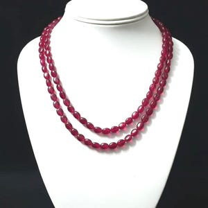 Op de afbeelding: Een ketting met twee strengen met ovale, dieprode kralen. De ketting wordt gepresenteerd op een witte sieraden buste tegen een zwarte achtergrond. De kralen hebben een gepolijste afwerking.