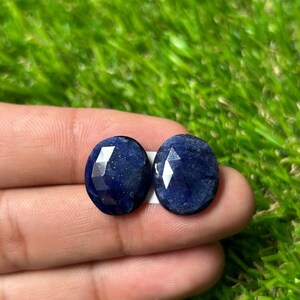 May include: Two oval-shaped, deep blue sapphires with a faceted surface texture. The gemstones exhibit a dark, saturated navy blue color with visible internal inclusions and a polished finish, reflecting light across their geometric cut surfaces.