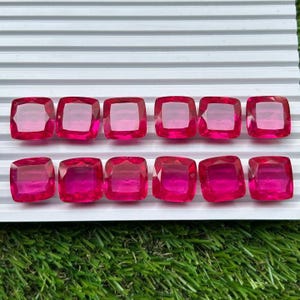 May include: Twelve loose cushion-cut gemstones in a vibrant hot pink hue. Each stone features a faceted surface that reflects light, arranged in two neat rows of six on a white grooved display tray. The stones are uniform in shape and color.