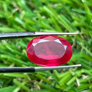 Könnte beinhalten: Ein oval geschliffener, leuchtend roter Edelstein, der von einer Metallpinzette gehalten wird. Der Stein hat eine facettierte Oberfläche, die das Licht reflektiert und einen tiefen karmesinroten Farbton sowie ein transparentes Aussehen zeigt.
