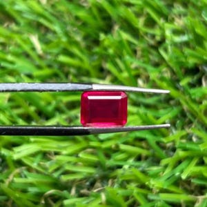 May include: A vibrant, rectangular-cut red gemstone held by a pair of black metal tweezers. The stone features a faceted surface that reflects light, showcasing a deep, saturated crimson hue throughout its transparent, polished crystalline structure.