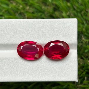 May include: Two oval-cut, deep red gemstones with faceted surfaces displayed side-by-side on a white rectangular jewellery display stand. The stones exhibit a vibrant, translucent crimson hue with light reflecting off the polished facets.