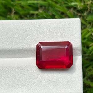 Pode incluir: Uma pedra preciosa de cor vermelho profundo e vibrante com um corte esmeralda retangular. A pedra apresenta facetas nítidas e limpas que refletem a luz na sua superfície polida, exibindo um tom carmesim rico e saturado.