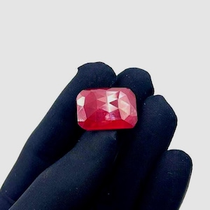 May include: A faceted, rectangular ruby gemstone. The gem is a vibrant red colour and is held by a gloved hand against a white background. The cut of the stone creates multiple reflective surfaces.