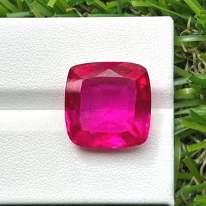 May include: A vibrant, cushion-cut gemstone with a deep, saturated fuchsia-pink color. The stone features a faceted surface that reflects light, creating a brilliant, sparkling appearance. It is positioned on a textured white surface.