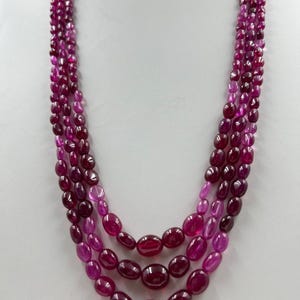 Op de afbeelding: Een meerstrengs ketting met ovale, dieprode edelstenen. De ketting heeft een trapsgewijs ontwerp, met grotere stenen in het midden en kleinere naar boven. De edelstenen hebben een gepolijste afwerking.