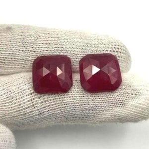 Puede incluir: Dos gemas facetadas, de corte cuadrado, de color rojo intenso. Las gemas tienen una superficie pulida y reflectante, sobre un fondo blanco. Las gemas son probablemente rubíes y son adecuadas para la fabricación de joyas.