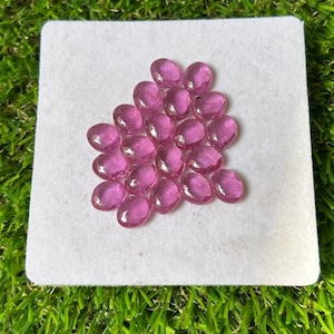 May include: A collection of small, oval-shaped, translucent pink gemstones arranged in a cluster on a square white card. Each stone features a smooth, polished surface with a rounded cabochon cut, displaying a consistent vibrant pink hue throughout.