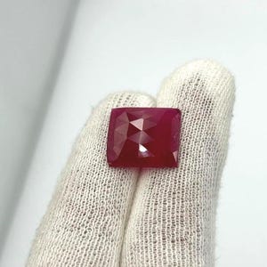 Op de afbeelding: Een gefacetteerde, vierkant geslepen robijn edelsteen. De robijn heeft een diepe, rijke rode kleur en meerdere facetten die licht reflecteren. De edelsteen wordt vastgehouden door een gehandschoende hand.