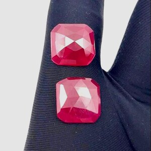 Peut inclure: Deux pierres précieuses rouge foncé, taillées en forme carrée. Les gemmes présentent une taille brillante avec de multiples facettes qui réfléchissent la lumière. Elles sont présentées sur un fond sombre, mettant en valeur leur couleur et leur éclat.