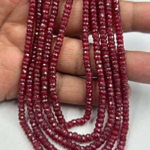 May include: Strands of faceted, deep red gemstone beads. The beads are small and uniform in size, strung together to create multiple necklaces. The color is a rich, dark red, with a slight sparkle from the facets. The beads are held in a hand.