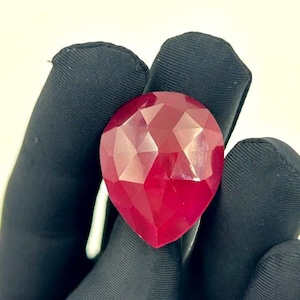 Puede incluir: Un anillo con una gema roja oscura facetada en forma de lágrima. La gema está sujeta por una banda plateada. El anillo se muestra sobre un fondo negro.