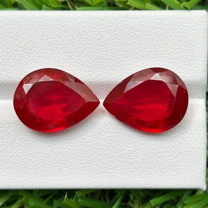 Può includere: Due gemme rosso vivo tagliate a forma di pera con molteplici sfaccettature. Le pietre sono posizionate fianco a fianco su una superficie espositiva bianca. Ogni gemma presenta una profonda tonalità cremisi e una superficie lucida e riflettente.