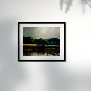 Puede incluir: Fotografía de paisaje enmarcada que presenta una masa de agua que refleja el cielo y la vegetación circundante. La escena incluye un campo de plantas doradas, árboles y un cielo nublado. La fotografía está en un marco negro.