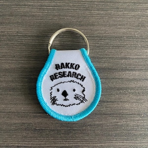 May include: A white and blue keychain with the text "RAKKO RESEARCH" and a cartoon otter face. The keychain has a silver key ring and a blue stitched border. The background is a wooden surface.