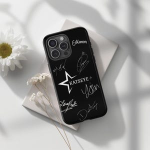 May include: Black phone case featuring multiple white signatures and the text "KATSEYE +" with a star graphic. The case has a camera module and is displayed on a white surface with floral accents.