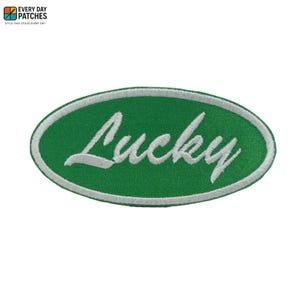 Könnte beinhalten: Grüner ovaler Aufnäher mit weiß gesticktem Text „Lucky“. Der Aufnäher hat einen weißen Rand und besteht aus einem strukturierten Stoff. Der Aufnäher ist ein schlichtes Design, das für Kleidung oder Accessoires geeignet ist.