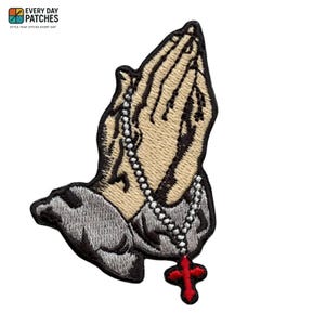 Peut inclure: Écusson brodé représentant des mains jointes en prière, un chapelet et une croix rouge. L'écusson est de couleur beige, grise et noire. L'écusson est de Every Day Patches.