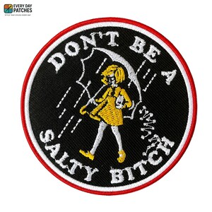 May include: Circular embroidered patch with a black background, a red border, and white text reading "DON'T BE A SALTY BITCH." It features a cartoon girl in a yellow dress under an umbrella in the rain.
