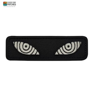 May include: A rectangular black embroidered patch featuring two stylised eyes with white concentric circular patterns. The design includes the text EVERY DAY PATCHES in the top left corner, presented in a clean, bold, and legible font style.