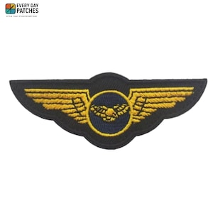 May include: Black embroidered patch with a gold-coloured wing design and a central emblem. The patch has a navy blue circular centre with a gold-coloured bird-like emblem. The patch is from Every Day Patches.