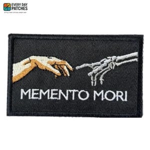 May include: Black rectangular patch featuring an embroidered design of two hands, one human and one skeletal, nearly touching. The words "MEMENTO MORI" are embroidered in white below the hands. The patch has a black border.