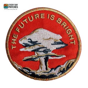 May include: Circular embroidered patch with the words "THE FUTURE IS BRIGHT" in gold thread on a red backdrop. The patch features a stylised mushroom cloud design in white, black, and gold. The patch is from Everyday Patches.