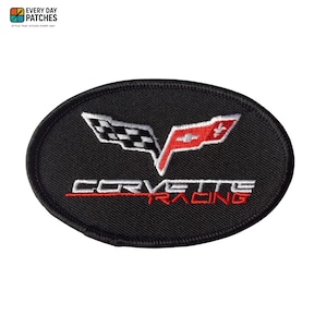 Op de afbeelding: Ovale zwarte patch met het Corvette Racing logo. Het logo toont een geblokte vlag en een rood-wit embleem, met de woorden "CORVETTE RACING" in wit en rood. De patch heeft een zwarte rand.