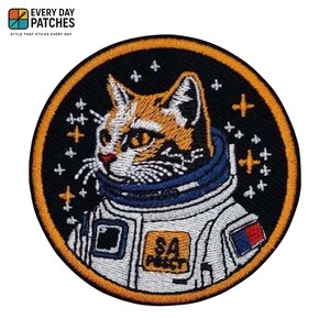 Könnte beinhalten: Runder, gestickter Aufnäher mit einer Katze in einem Astronautenanzug. Die Katze hat orange-weißes Fell, einen blauen Kragen und einen weißen Raumanzug. Der Aufnäher hat einen schwarzen Hintergrund mit Sternen und dem Text "SA PUSSY".