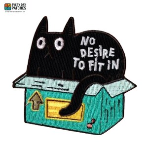 Puede incluir: Parche bordado con un gato negro dentro de una caja turquesa. El gato tiene ojos blancos y el texto "NO DESIRE TO FIT IN" está encima. La caja tiene una flecha y una etiqueta amarilla.