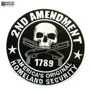 Könnte beinhalten: Runder schwarzer Aufnäher mit weißem Text und einer Totenkopf-Grafik. Der Text lautet "2ND AMENDMENT" und "AMERICA'S ORIGINAL HOMELAND SECURITY" mit dem Jahr "1789". Zwei gekreuzte Pistolen befinden sich unter dem Totenkopf.